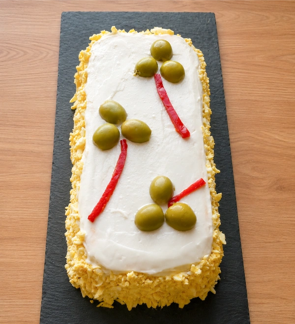 Vista cenital de un pastel de atún con pan de molde decorado con mayonesa, aceitunas rellenas, piquillos y borde de huevo rallado