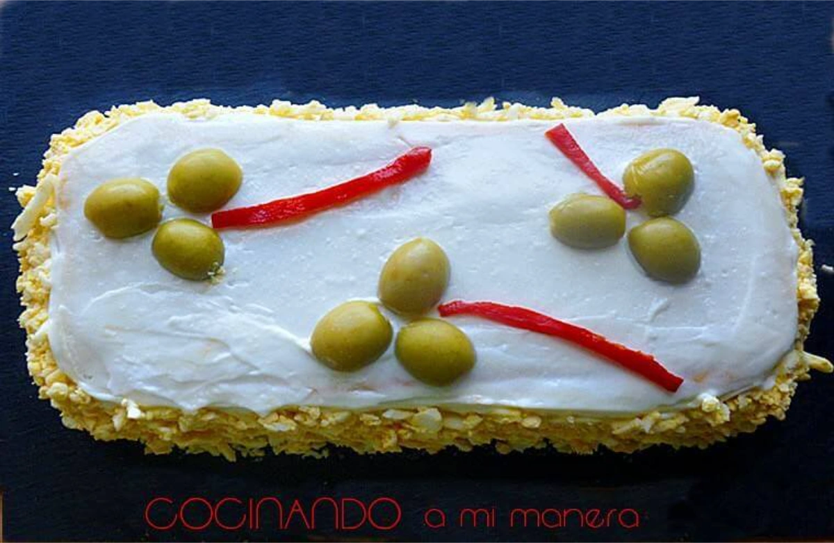 Pastel de atún frío con pan de molde cubierto de mayonesa, con huevo cocido rallado en los laterales y decorado con aceitunas y tiras de piquillo