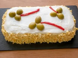 Pastel de atún frío con pan de molde sobre tabla de pizarra, cubierto de mayonesa y huevo cocido rallado, decorado con aceitunas y tiras de piquillo