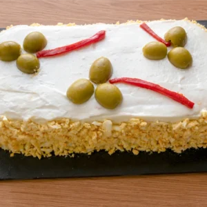 Pastel de atún frío con pan de molde sobre tabla de pizarra, cubierto de mayonesa y huevo cocido rallado, decorado con aceitunas y tiras de piquillo