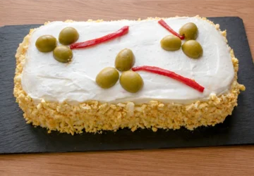Pastel de atún frío con pan de molde sobre tabla de pizarra, cubierto de mayonesa y huevo cocido rallado, decorado con aceitunas y tiras de piquillo
