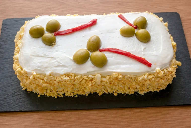 Pastel de atún frío con pan de molde sobre tabla de pizarra, cubierto de mayonesa y huevo cocido rallado, decorado con aceitunas y tiras de piquillo