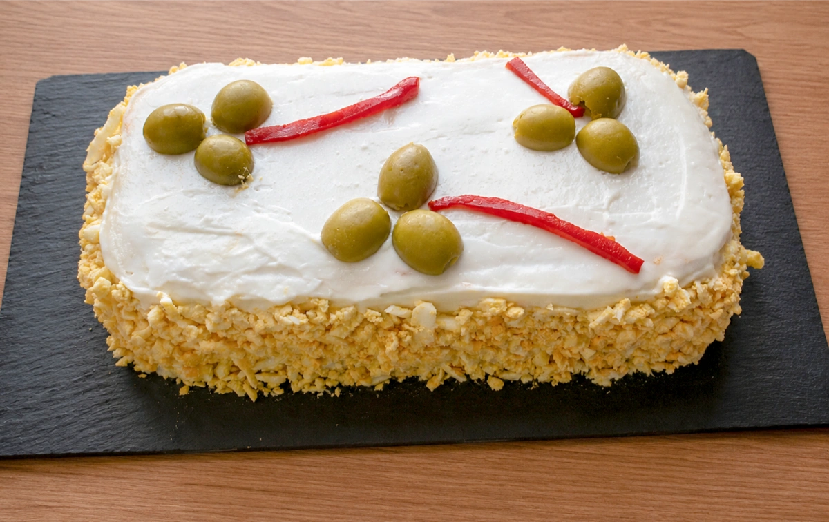 Pastel de atún frío con pan de molde sobre tabla de pizarra, cubierto de mayonesa y huevo cocido rallado, decorado con aceitunas y tiras de piquillo