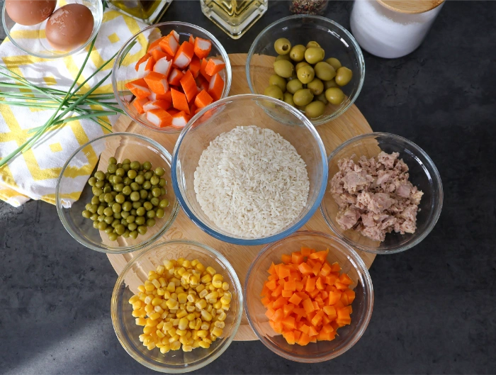 Ingredientes de la ensalada de arroz: arroz, atún, guisantes, maíz, zanahoria, surimi y aceitunas en cuencos