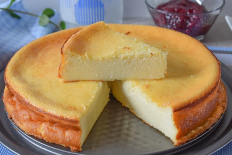 Tarta de queso al horno con una porción levantada mostrando el interior cremoso y cuenco de mermelada de frambuesa al fondo
