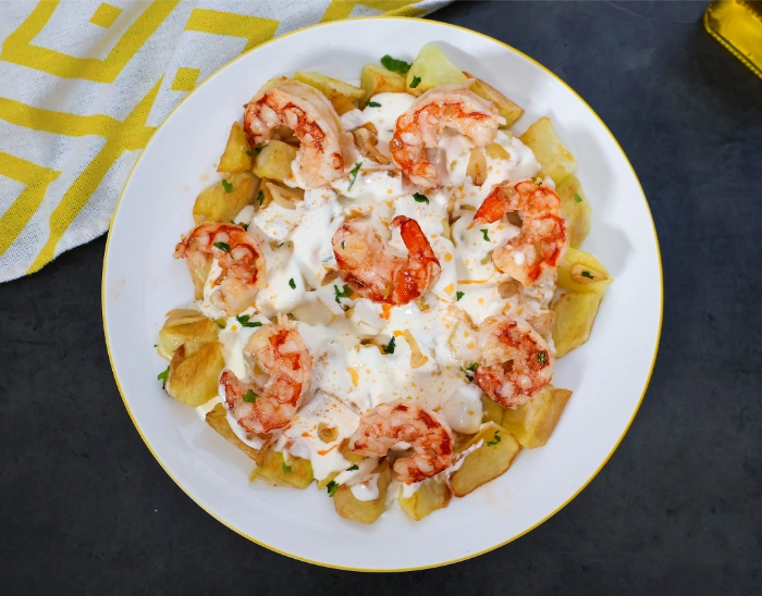 Patatas arrieras con gambones y alioli casero servidas en plato blanco con borde dorado, vistas desde arriba