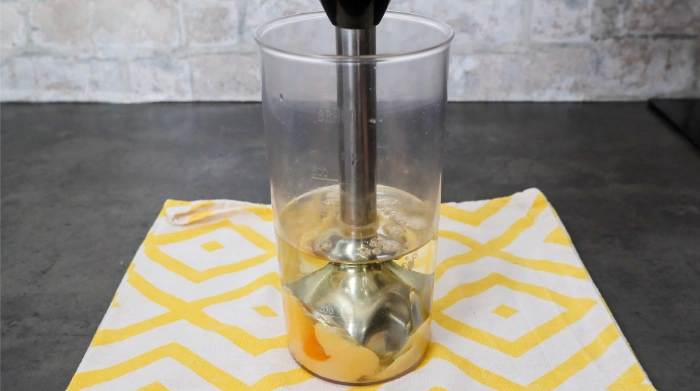 Preparación de alioli casero con batidora de mano en vaso de cristal alto, con huevo y aceite de oliva visibles en el fondo