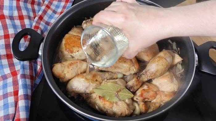 Mano vertiendo vino blanco sobre el pollo dorado con cebolla y laurel.