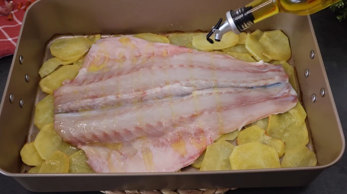 Lubina abierta colocada sobre patatas panadera, lista para hornear con aceite de oliva.