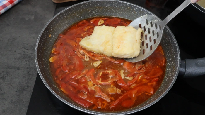 Lomo de bacalao marcado incorporándose con una espumadera a la sartén con ajada roja y pimientos asados,