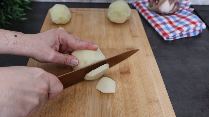 Manos cortando patatas en rodajas de grosor medio sobre tabla de madera, preparación del acompañamiento del bacalao a la tranca