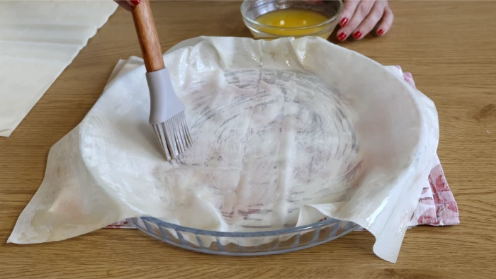 Pincelando una lámina de masa filo con mantequilla y aceite en fuente redonda para montar la kotopita