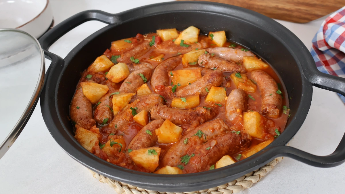 Cazuela de hierro con salchichas en salsa de tomate y patatas, paño de cuadros y tapa de cristal, receta casera terminada