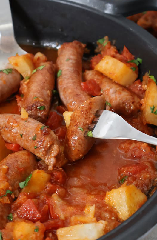 Tenedor pinchando una salchicha jugosa en salsa de tomate con patatas y perejil fresco, formato vertical