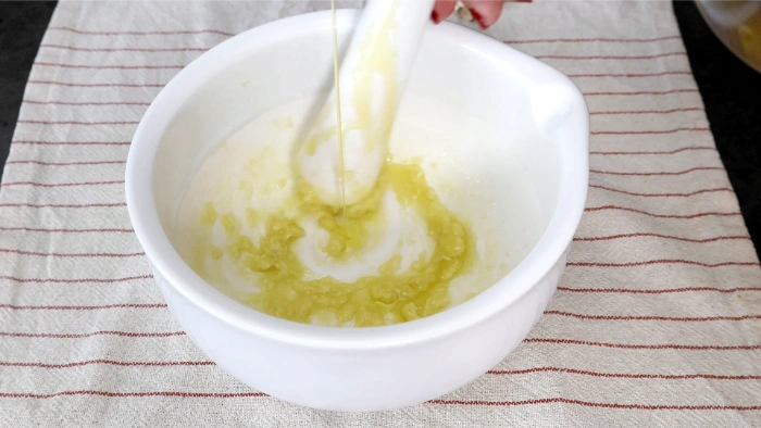 Hilo fino de aceite de oliva virgen extra cayendo sobre la mezcla de pan y ajo en bol blanco, formando la emulsión del aliño del cojondongo extremeño