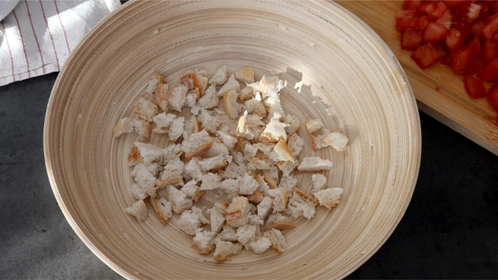 Bol de madera con pan de corteza troceado en la base, primer paso del montaje del cojondongo extremeño, al fondo tomate picado sobre tabla