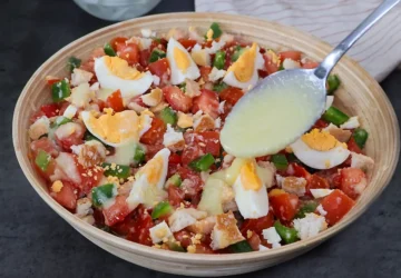 Cuchara vertiendo aliño cremoso de pan y ajo sobre cojondongo extremeño terminado con tomate, pimiento y huevo cocido en cuartos, listo para servir