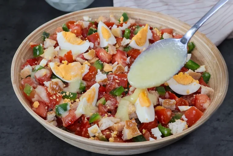 Cuchara vertiendo aliño cremoso de pan y ajo sobre cojondongo extremeño terminado con tomate, pimiento y huevo cocido en cuartos, listo para servir