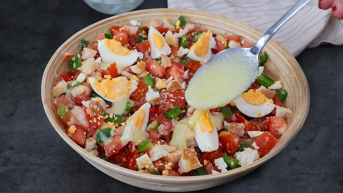 Cuchara vertiendo aliño cremoso de pan y ajo sobre cojondongo extremeño terminado con tomate, pimiento y huevo cocido en cuartos, listo para servir