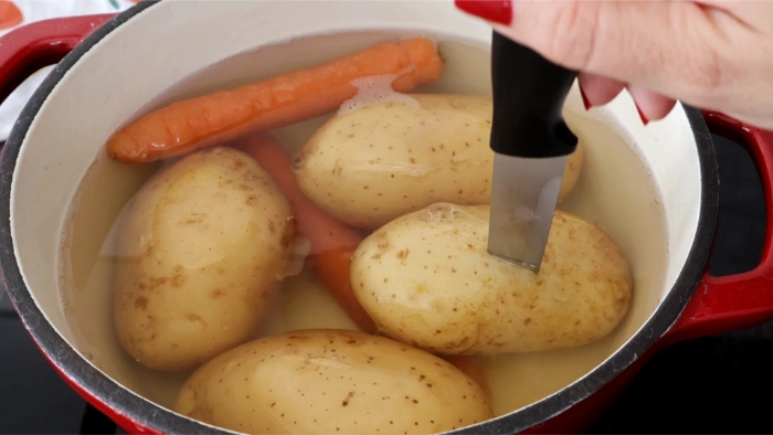 Patatas y zanahoria enteras con piel cociéndose en una cazuela, comprobando el punto de cocción con un cuchillo