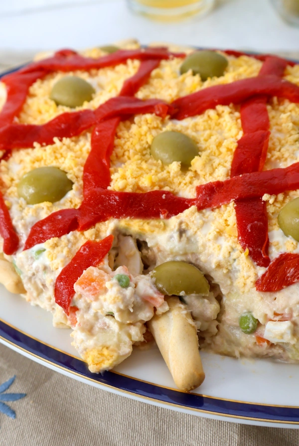 Primer plano de ensaladilla rusa cremosa con guisantes, zanahoria y huevo, decorada con pimiento, yema y aceitunas