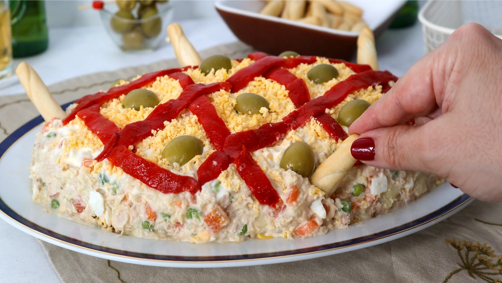 Mano cogiendo un colín para mojar en la ensaladilla rusa decorada con pimiento, yema y aceitunas