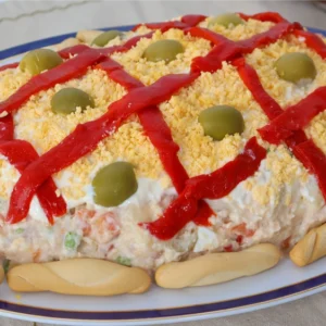 Ensaladilla rusa casera decorada con enrejado de pimiento rojo, yema de huevo desmenuzada, aceitunas y colines alrededor