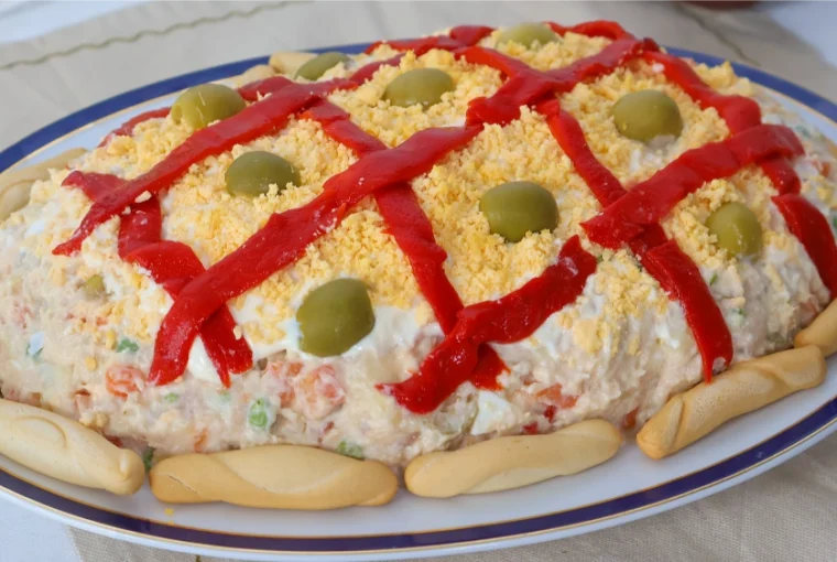 Ensaladilla rusa casera decorada con enrejado de pimiento rojo, yema de huevo desmenuzada, aceitunas y colines alrededor