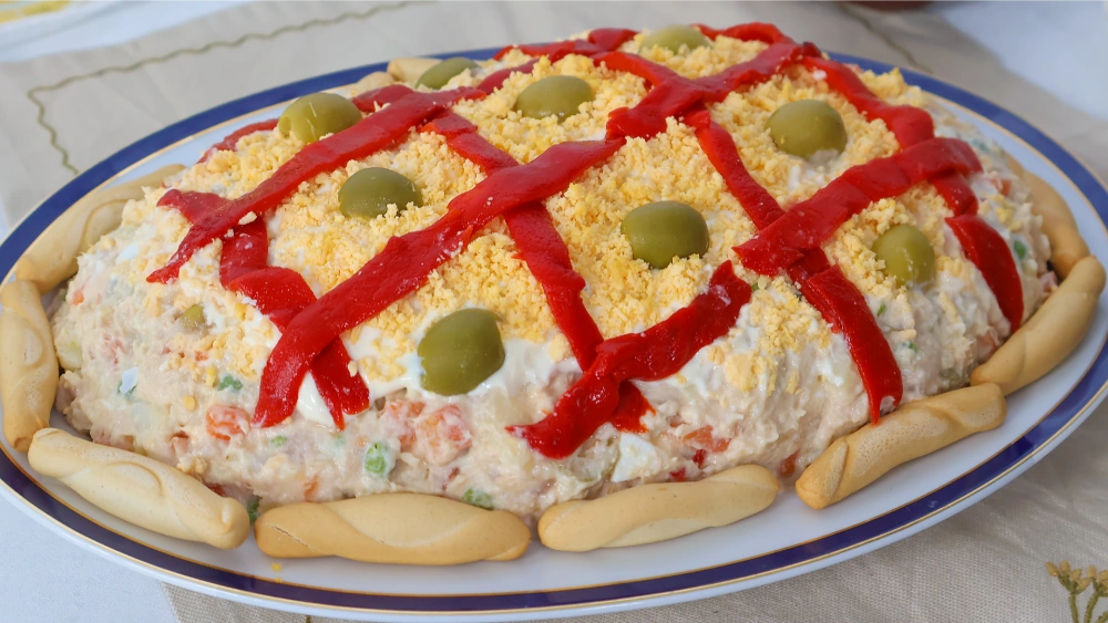 Ensaladilla rusa casera decorada con enrejado de pimiento rojo, yema de huevo desmenuzada, aceitunas y colines alrededor