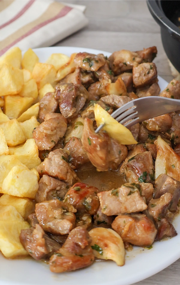 Imagen vertical mostrando trocito de carne al ajillo en tenedor junto con patata frita