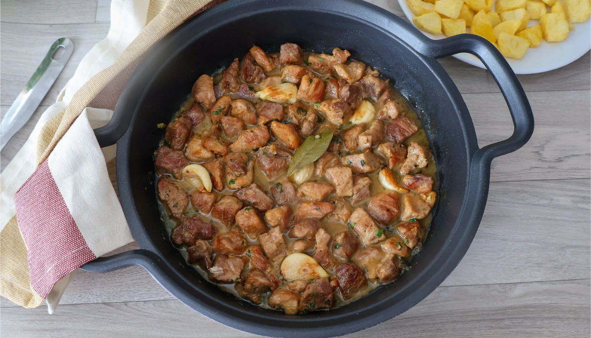 Cazuela con carne de cerdo al ajillo recién cocinada y servida junto a plato de patatas